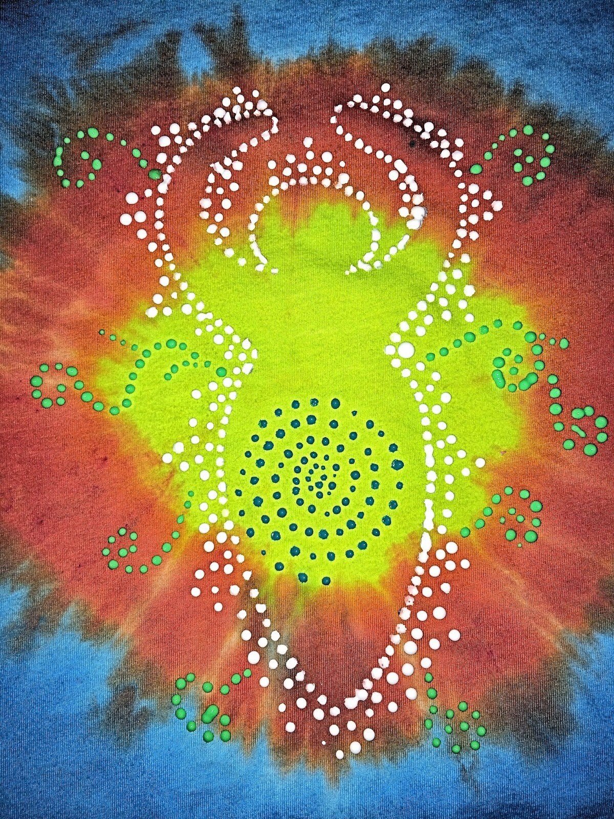 Close-up of the finished tie-dye goddess shirt showing the dot-painted goddess silhouette with white and green puffy paint dots and a blue spiral center on a vibrant sunburst background